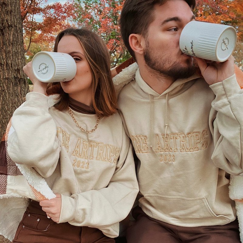 Customer photo: couple in matching cream embroidered sweatshirts sipping from mugs under autumn trees